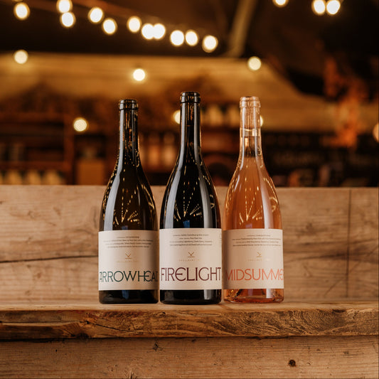 Three wine bottles from the brand Arrowweaver, labeled Arrowhead, Firelight, and Midsummer, displayed on a rustic wooden shelf against a warm, softly blurred background with hanging string lights.