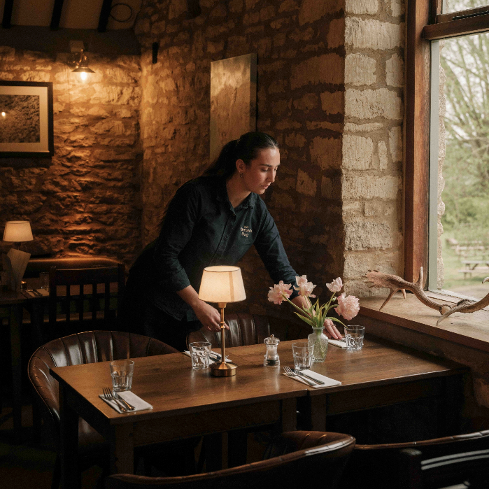 Woman arranging flowers on a table at The Farmer'ss Dog Pub in a cozy setting with stone walls and large windows.